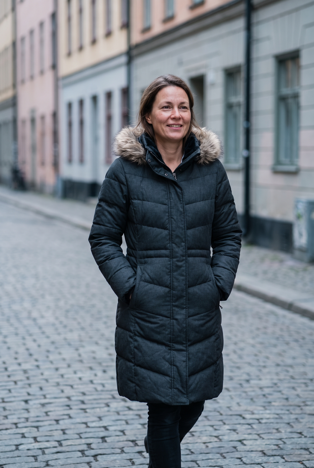 Kvinna i lång mörkgrå vadderad vinterjacka med pälskantad huva på en kullerstensgata i Stockholm