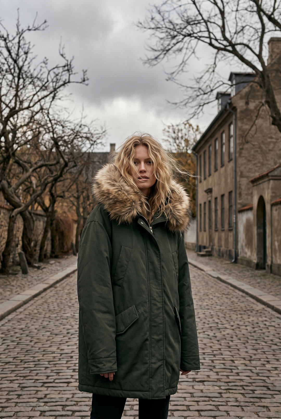 Kvinna i lång mörkgrön parkas med pälskantad huva på en blåsig höstgata