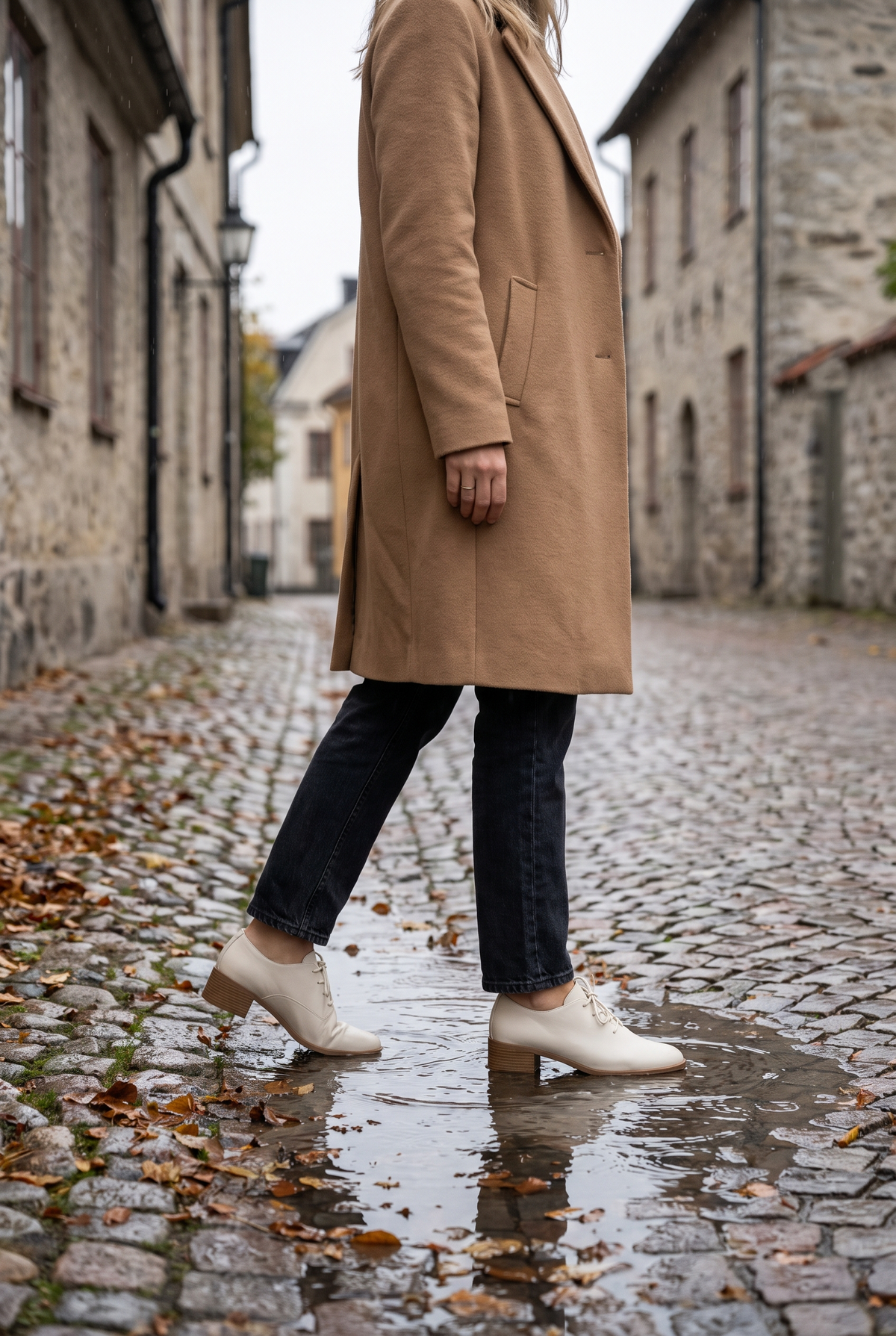 Kvinna i beige kappa och vattentäta skor som går över en blöt kullerstensgata en regnig höstdag