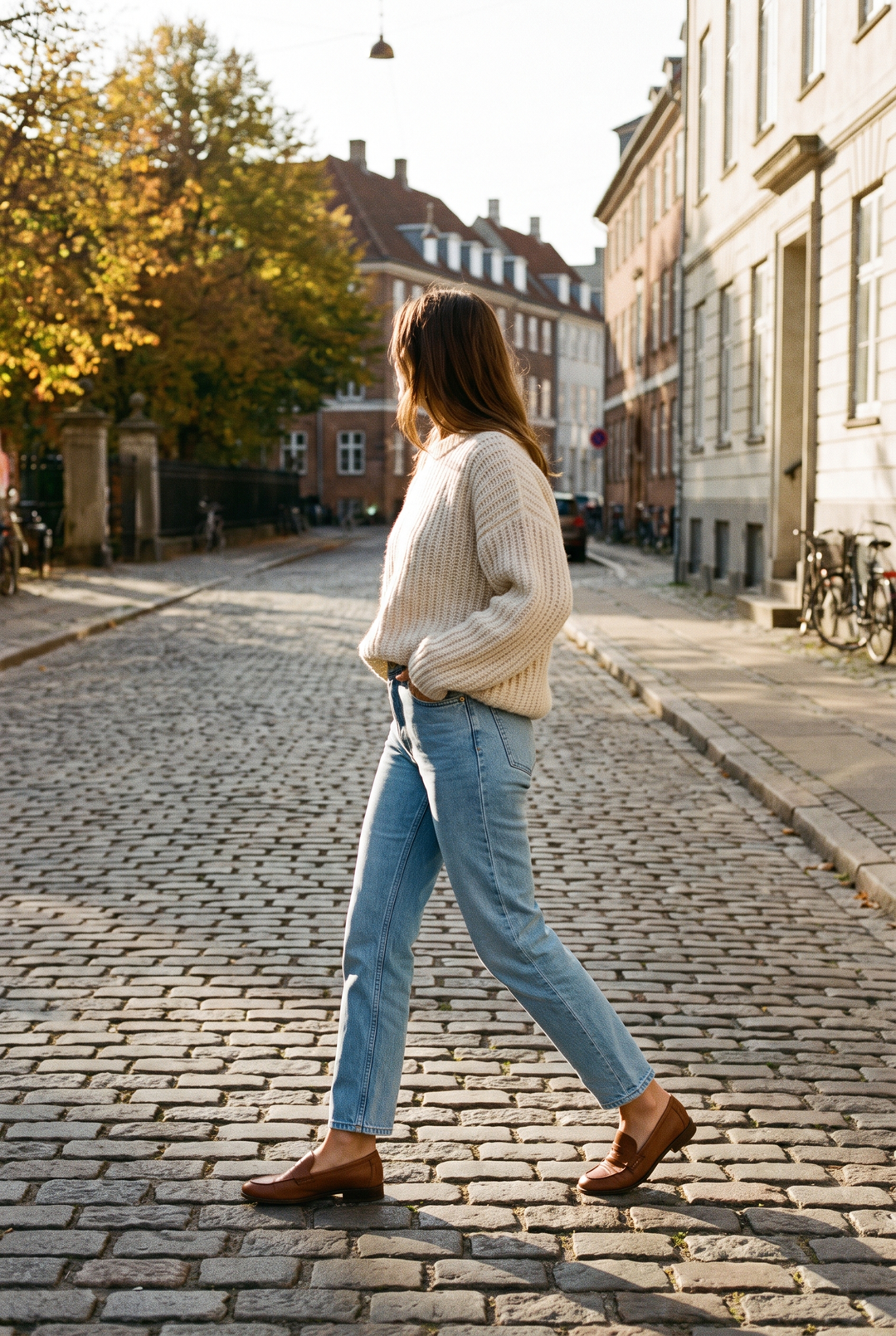 Kvinna i ljusblå raka jeans, cremevit stickad tröja och bruna loafers som går på en kullerstensgata i höstsol, jeansen slutar precis vid vristen