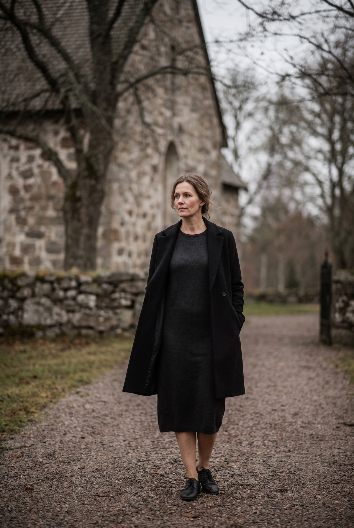 Kvinna i mörk långärmad klänning och svart lång kappa på en grusgång utanför en kyrka en grå höstdag