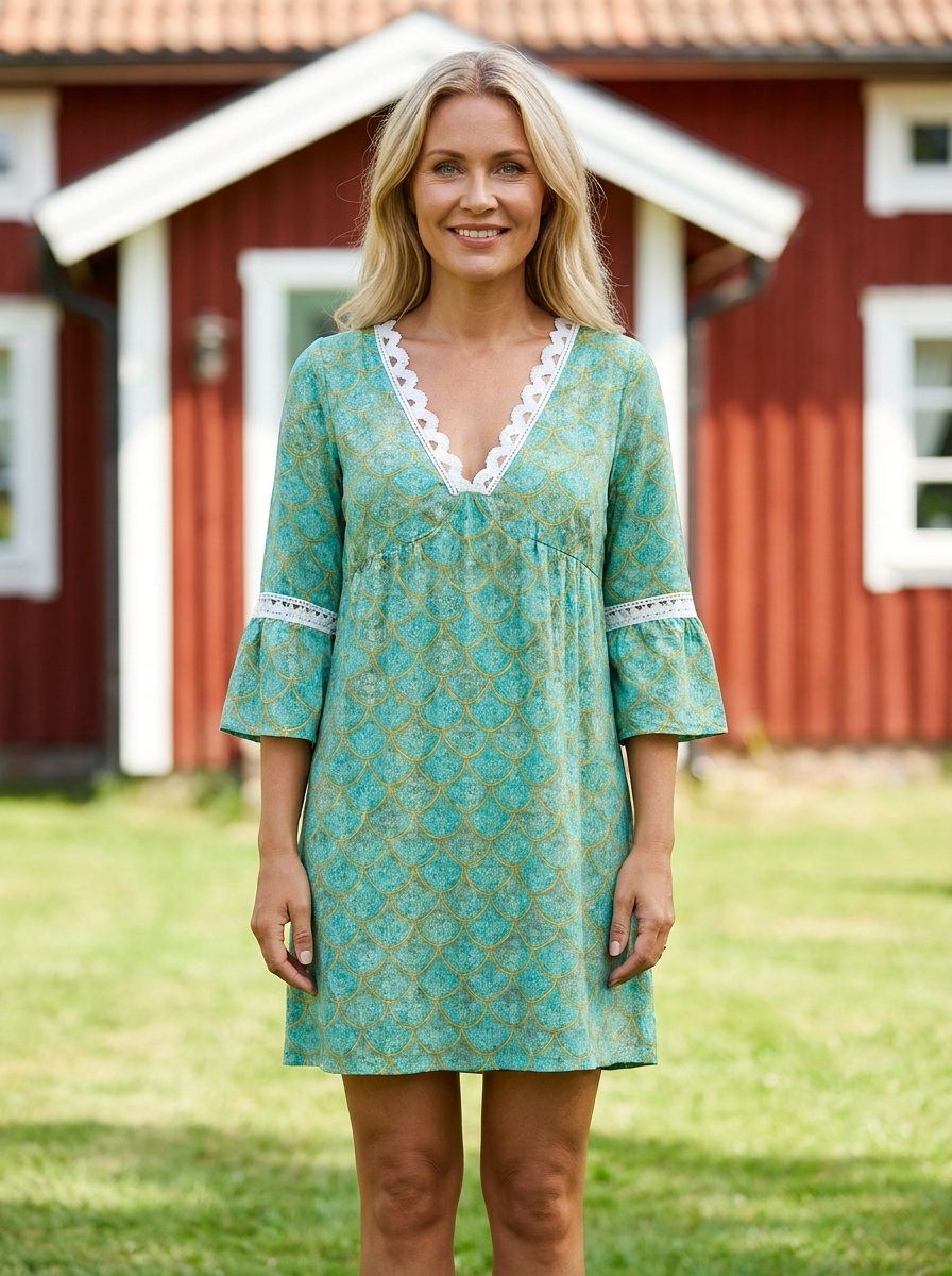 Alva knälång sommarklänning i turkosgrönt mönster med vit spetskant, buren framför rött sommarhus – Lissandra.se