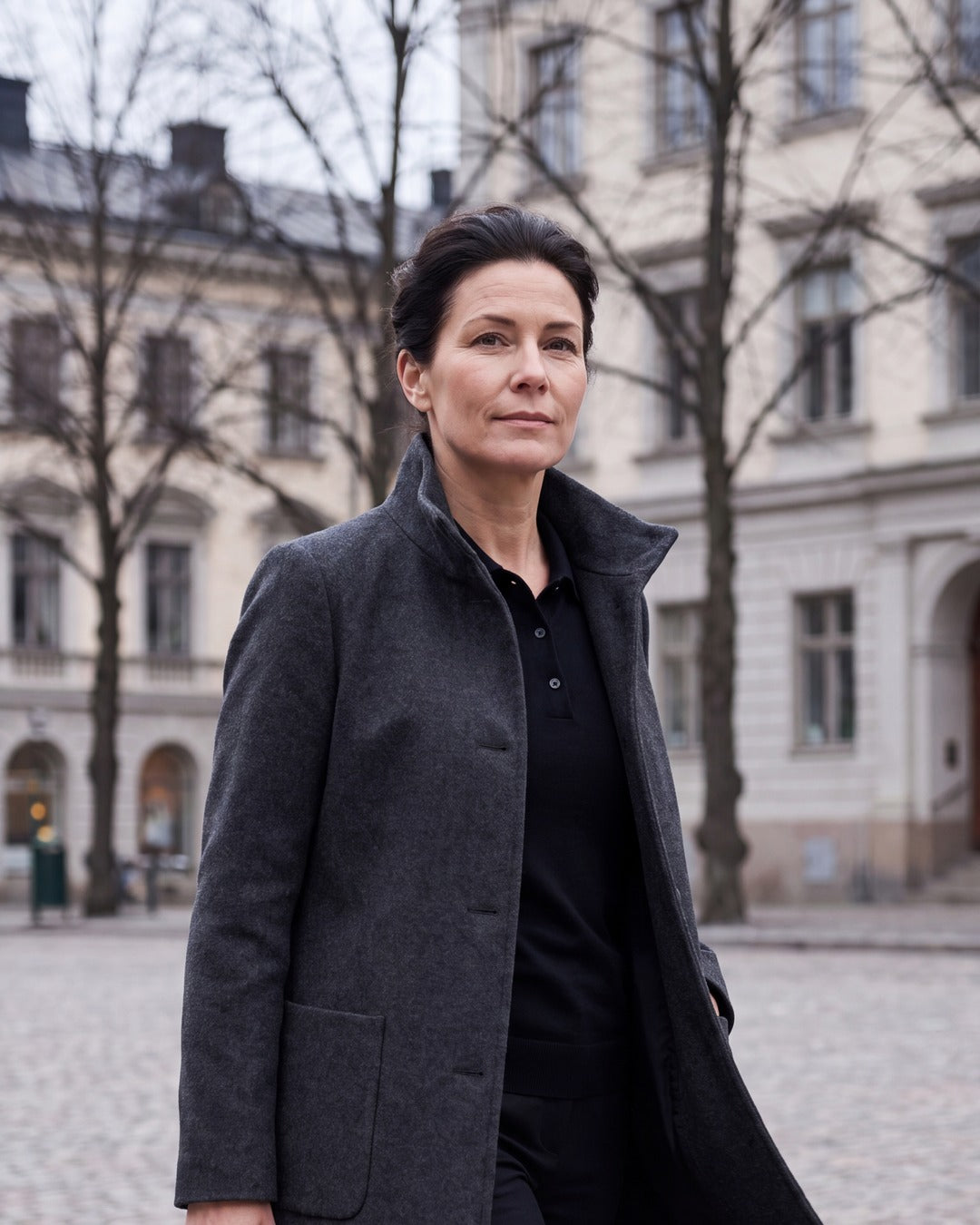Kvinna i mörkgrå ullkappa med hög ståkrage uppfälld, på ett stenlagt torg i Stockholm