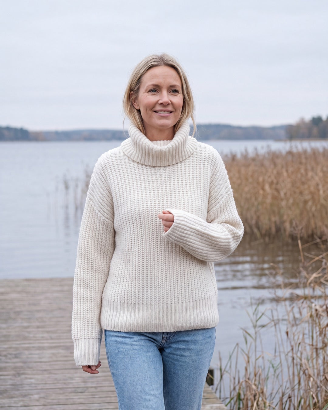 Kvinna i tjock vit stickad tröja med mjuk rullkrage på en brygga vid en svensk sjö om hösten