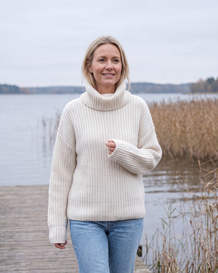 Kvinna i tjock vit stickad tröja med mjuk rullkrage på en brygga vid en svensk sjö om hösten