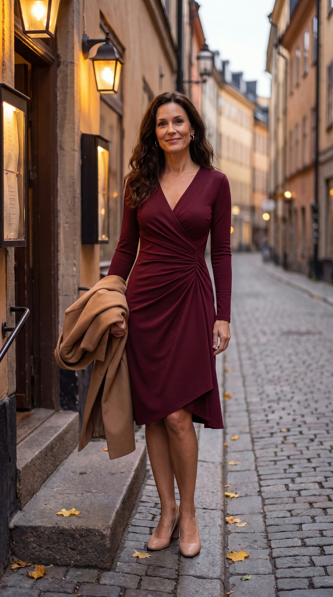 Kvinna i vinröd draperad midiklänning med V-ringning och långa ärmar på en kullerstensgata i höstkväll